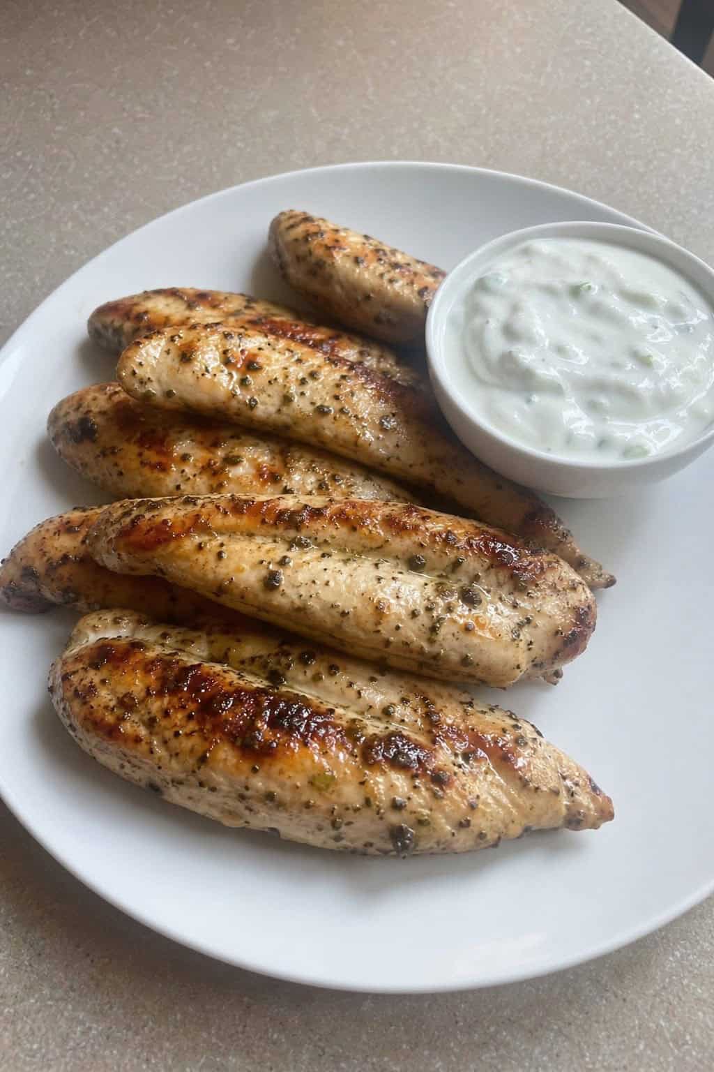 Herb-flecked chicken tenders on a white plate with a small bowl of dipping sauce and fresh parsley scattered on a wooden table.