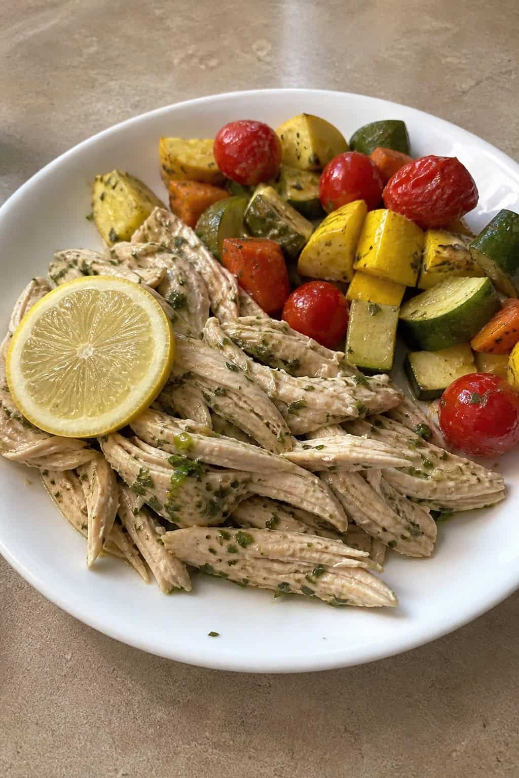 A white plate on a wooden table with lemon-herb shredded chicken resting alongside caramelized roasted carrots, zucchini coins, strips of red bell pepper, and blistered cherry tomatoes, with a sprinkle of fresh flat-leaf parsley.