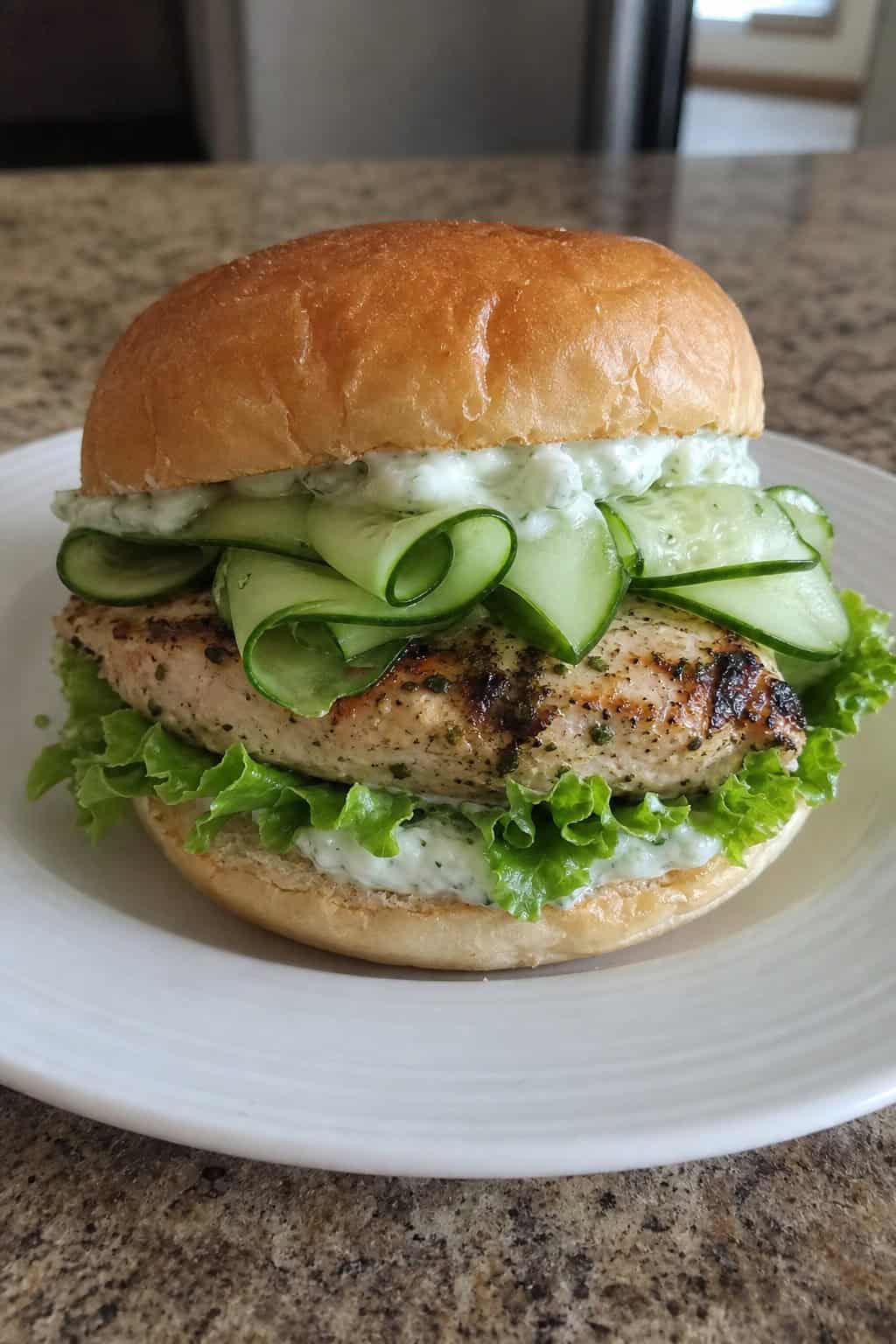 A grilled chicken sandwich with long cucumber ribbons, green lettuce, and a creamy sauce on a toasted roll, served on a wooden board with lemon wedges beside it