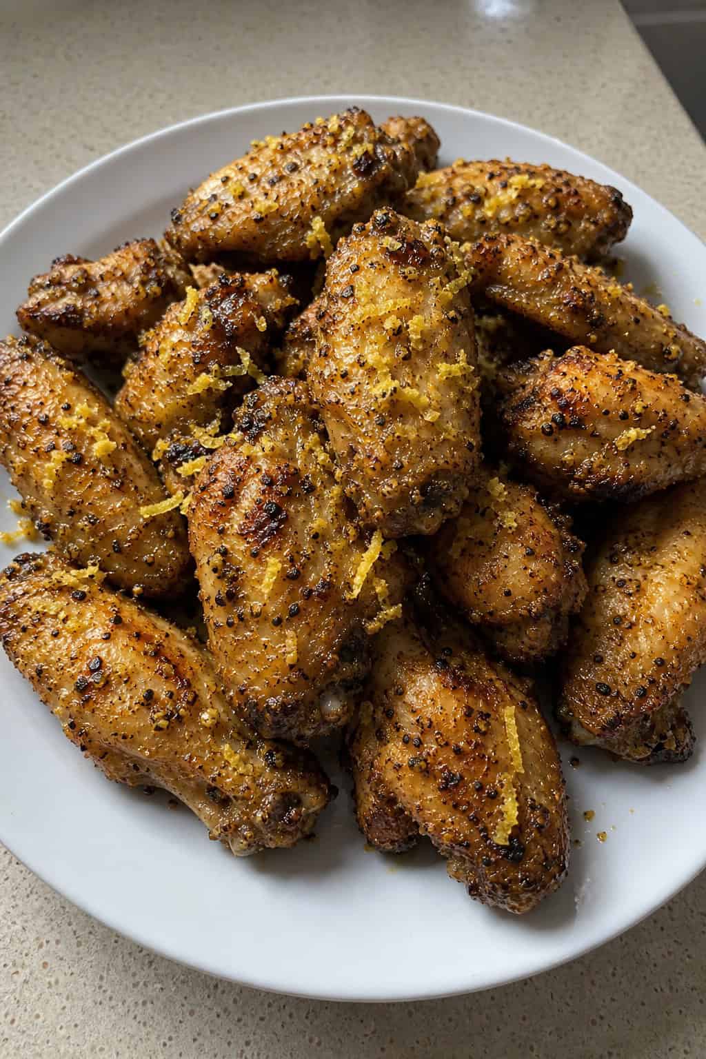A plate of golden brown lemon pepper chicken wings garnished with lemon zest and lemon slices, with a small bowl of dipping sauce and fresh herbs in the background.