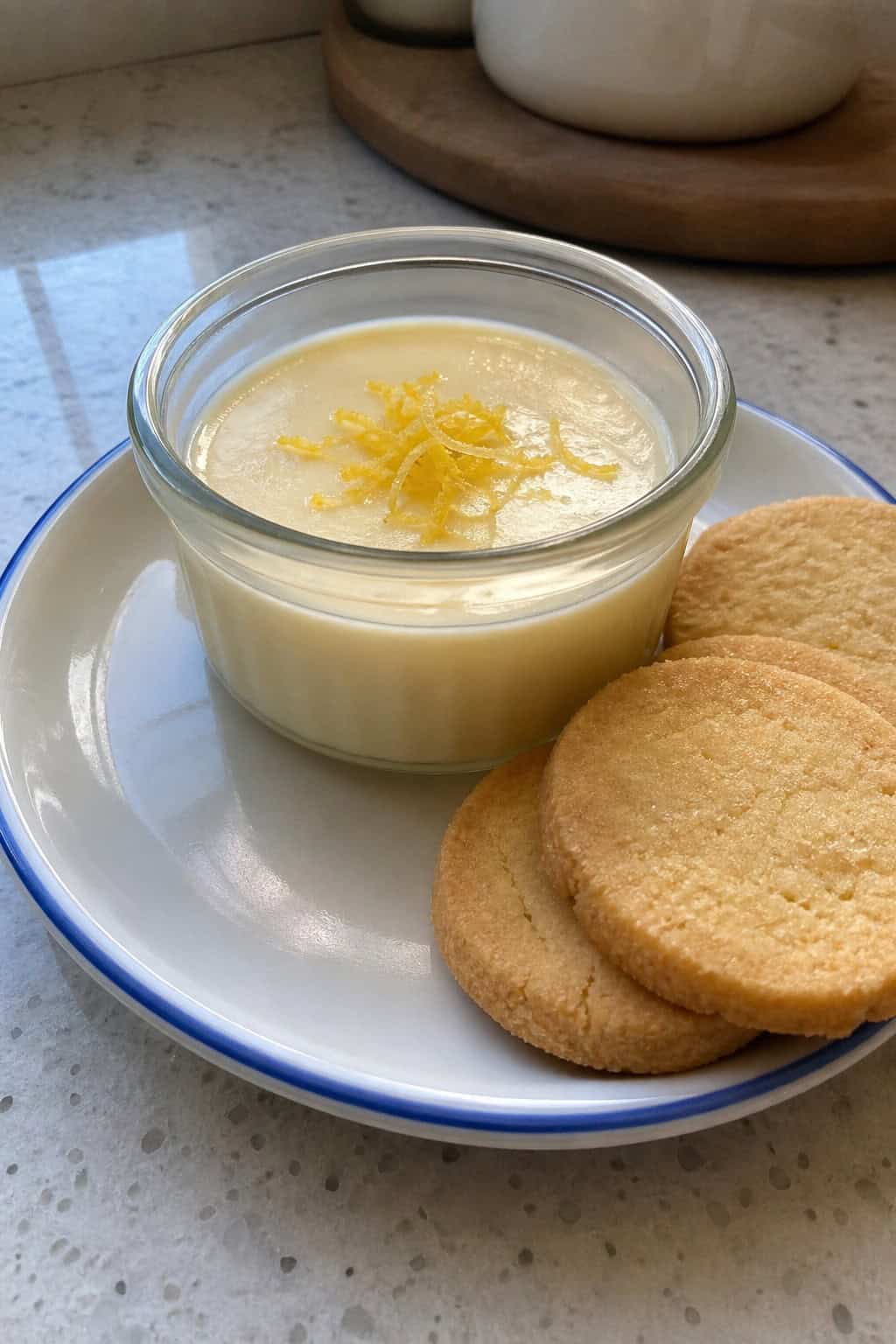 A lemon posset dessert in a glass ramekin with lemon zest on top, served alongside golden shortbread cookies on a wooden surface.