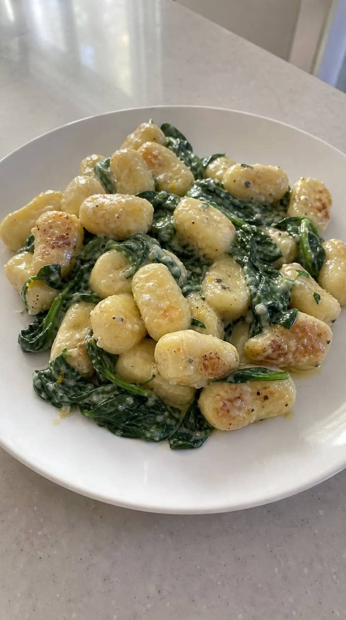 Plump gnocchi coated in a pale, creamy ricotta sauce with deep green wilted spinach leaves and thin curls of lemon zest scattered across a wide white bowl.
