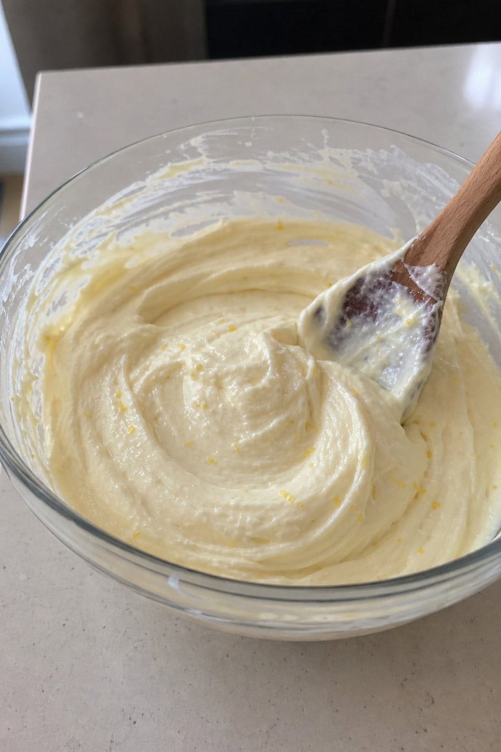 A glass bowl filled with creamy lemon-zest cheesecake filling surrounded by fresh lemons and a wooden spoon on a kitchen countertop.