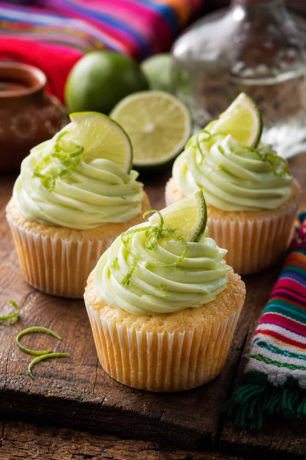 Three lime and tequila tres leches cupcakes with green frosting and lime garnishes on a wooden table.