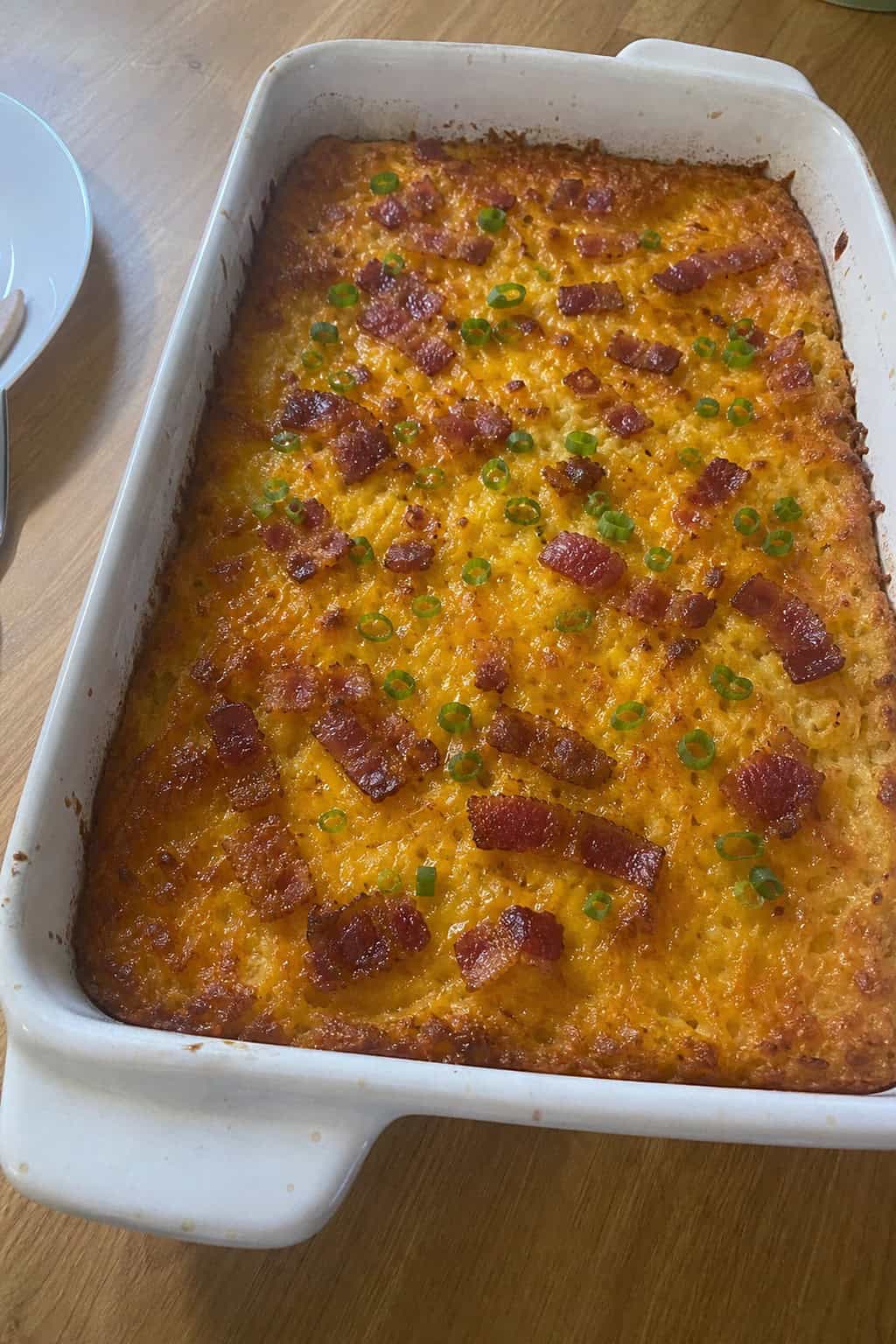 A baked potato hashbrown casserole topped with bacon and green onions in a baking dish on a wooden table.