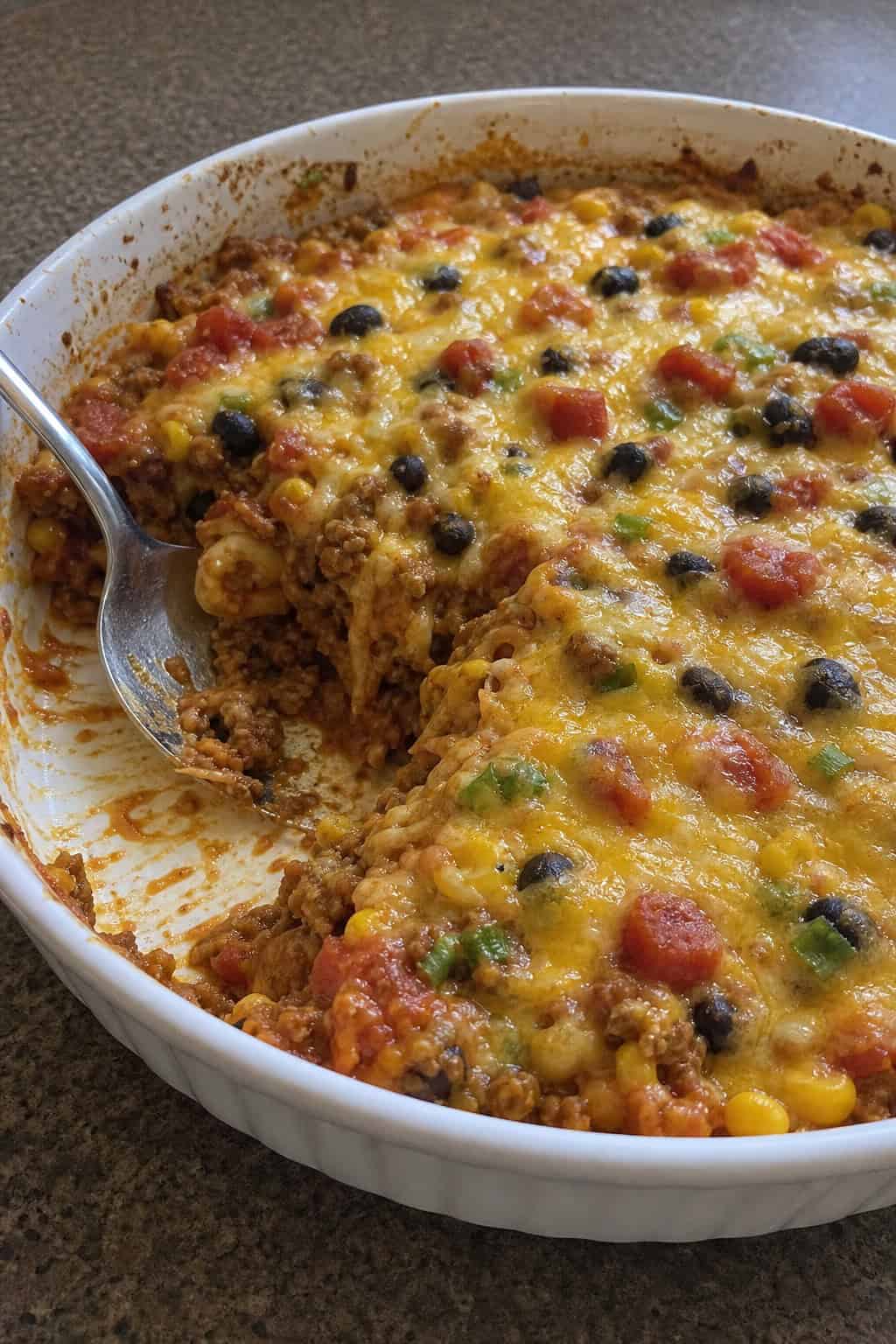 A baked taco-enchilada casserole with ground turkey in a ceramic dish, topped with melted cheese, corn kernels, black beans, and sliced green onions, with visible golden-brown edges around the casserole.