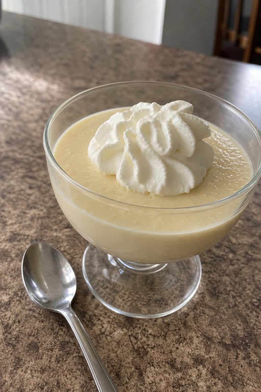 A glass cup filled with creamy cheesecake pudding topped with whipped cream and a mint leaf on a kitchen countertop with ingredients in the background.