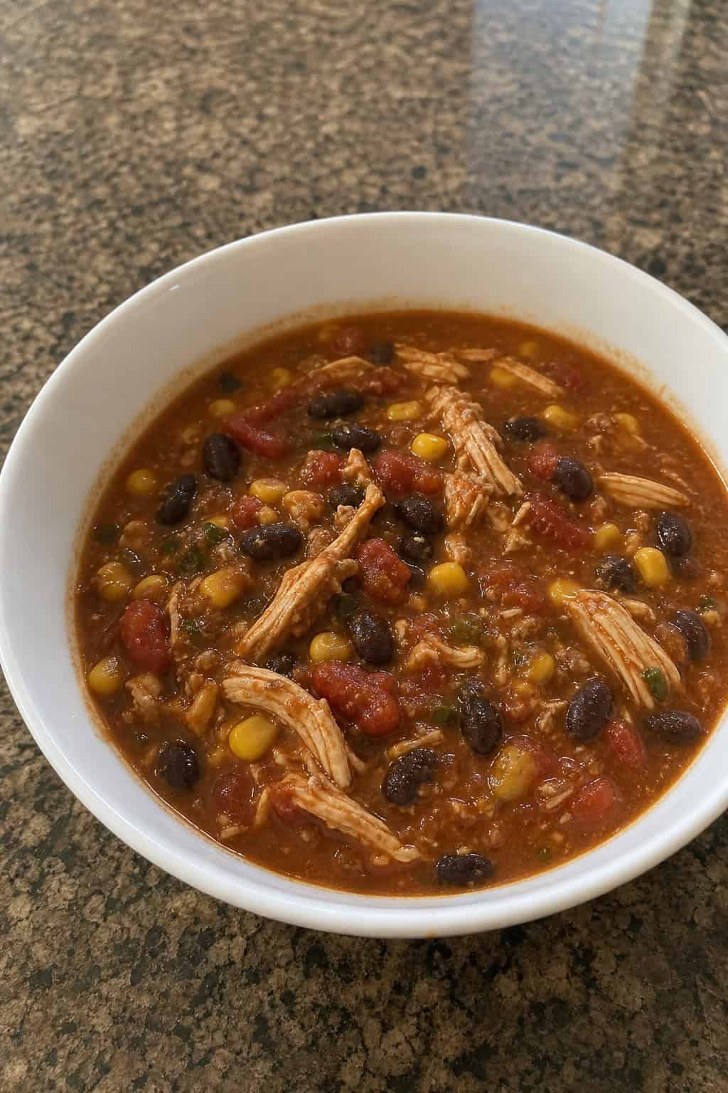 A bowl of chicken chili with black beans and vegetables on a wooden table with fresh ingredients around it.