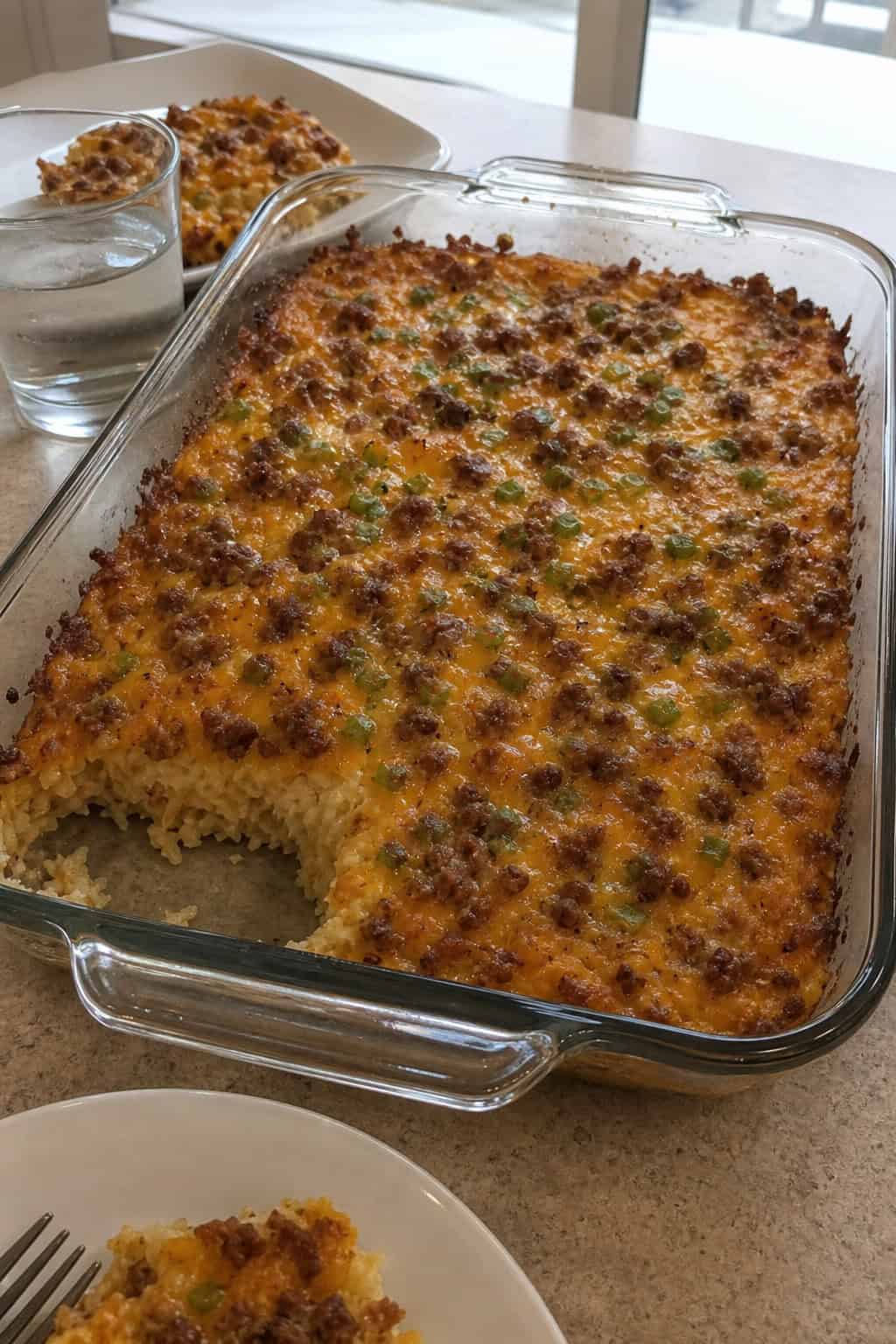 A family-size hashbrown casserole with sausage, cheese, and eggs in a foil baking pan on a wood table, with a glass baking dish and scattered fresh ingredients nearby.