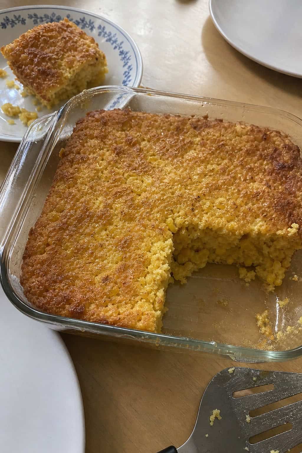 A golden-brown corn casserole in a clear glass baking dish on a wooden table with small decorative gourds and dried leaves arranged around it.