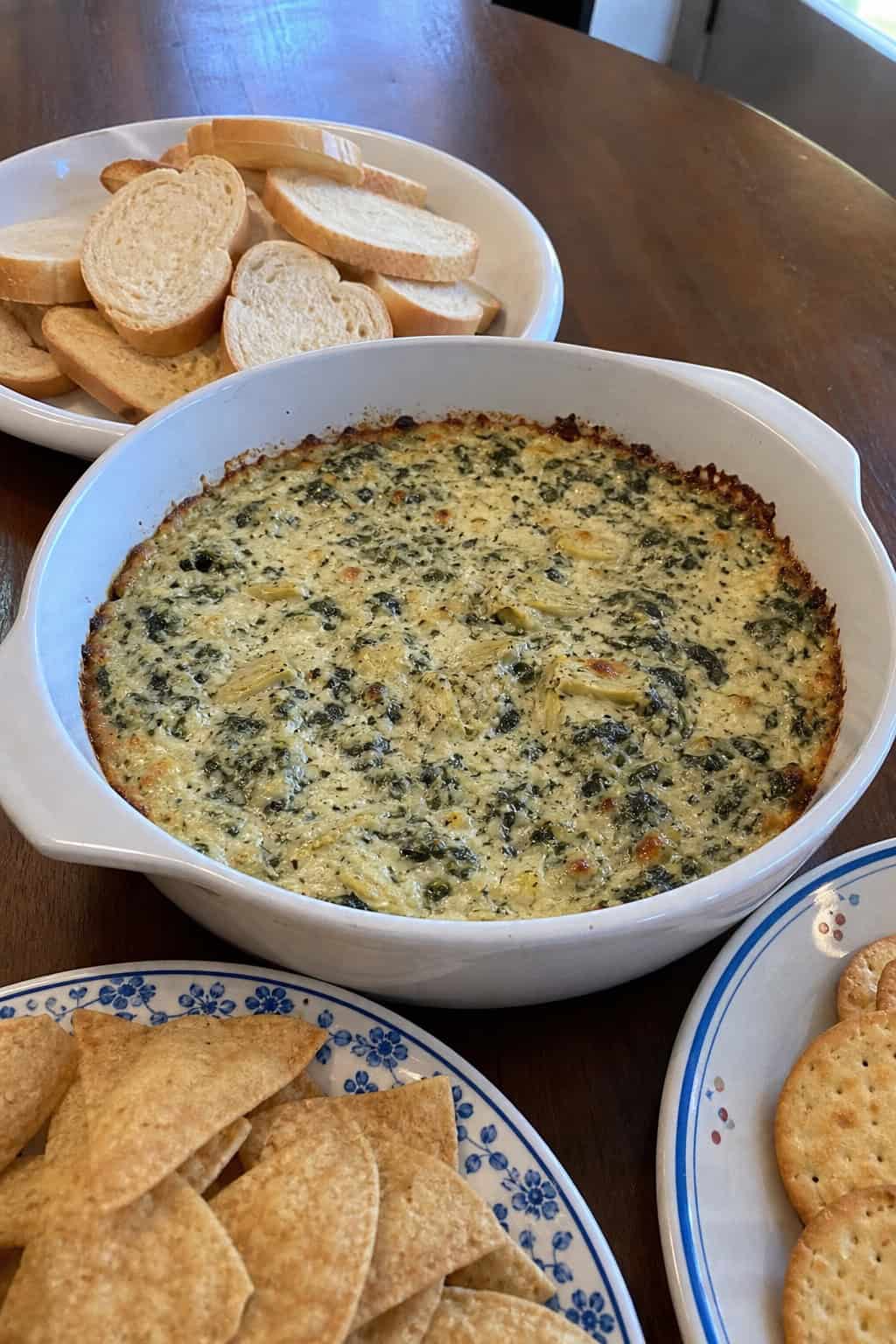 A baked spinach and artichoke dip in a white dish surrounded by bread slices and vegetable sticks on a wooden table.