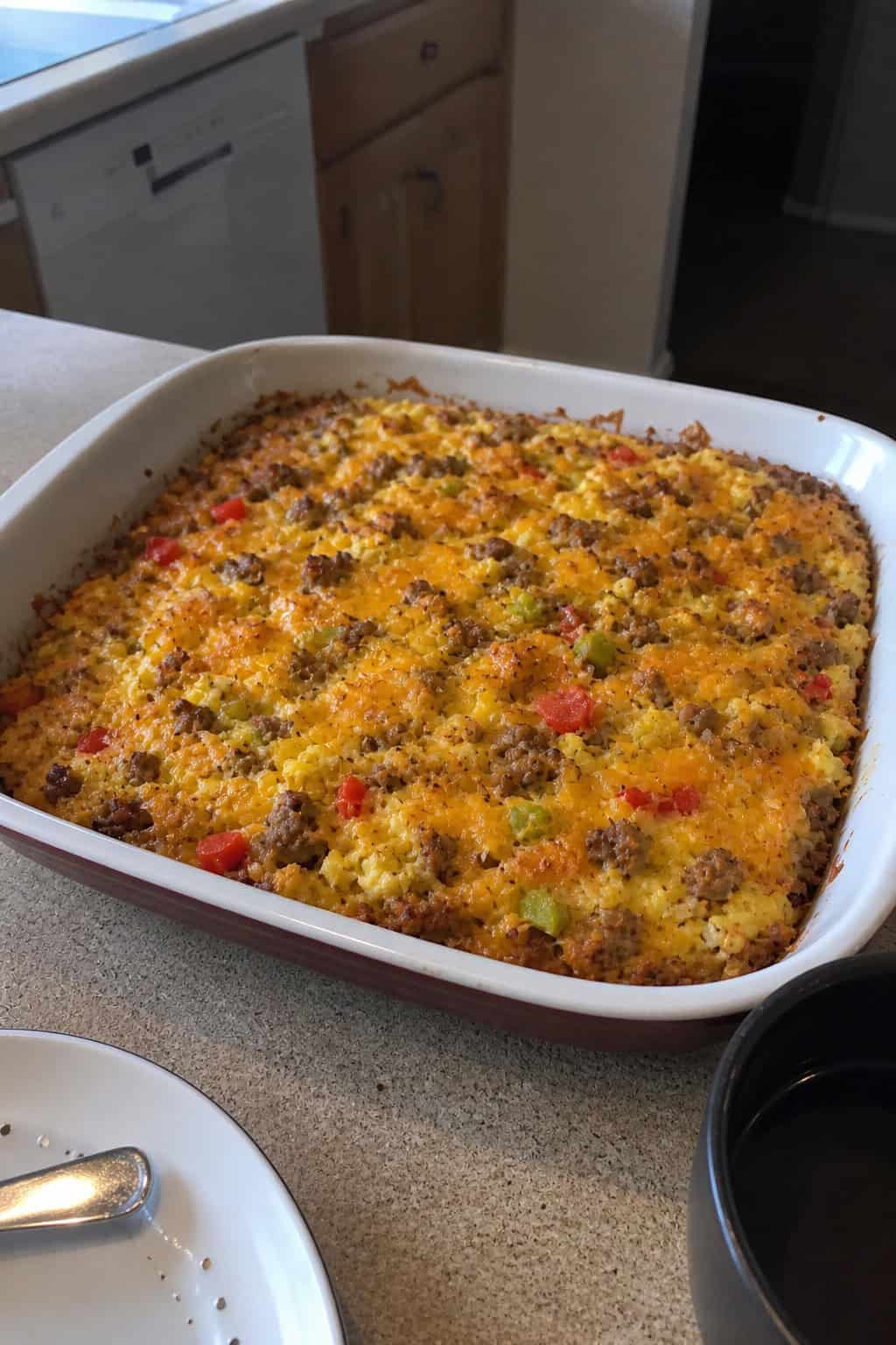 A hashbrown breakfast casserole in a glass baking dish on a wood table, surrounded by loose eggs, a block of cheese, a package of sausage, and a bag of frozen hashbrowns.