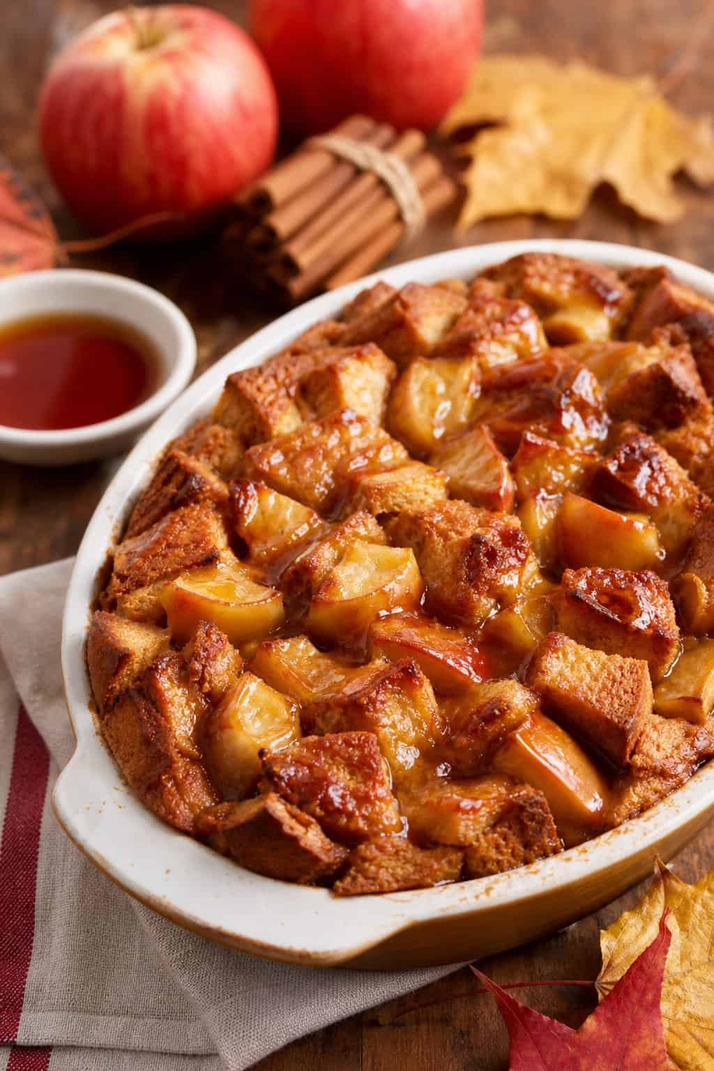 A plate of maple apple bread pudding with apples and cinnamon sticks on a wooden table.
