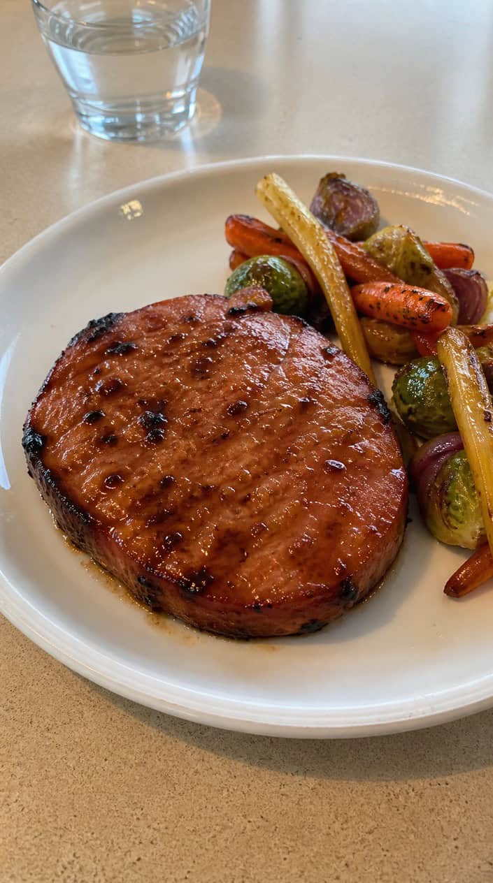 A glazed ham steak on a plate garnished with herbs and served with roasted vegetables on a kitchen table.