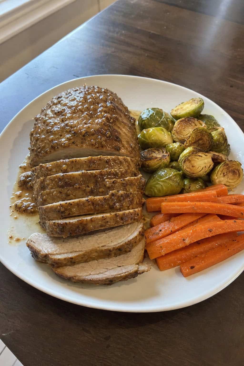 Sliced maple Dijon pork loin on a white plate with roasted vegetables and fresh herbs on a wooden table.