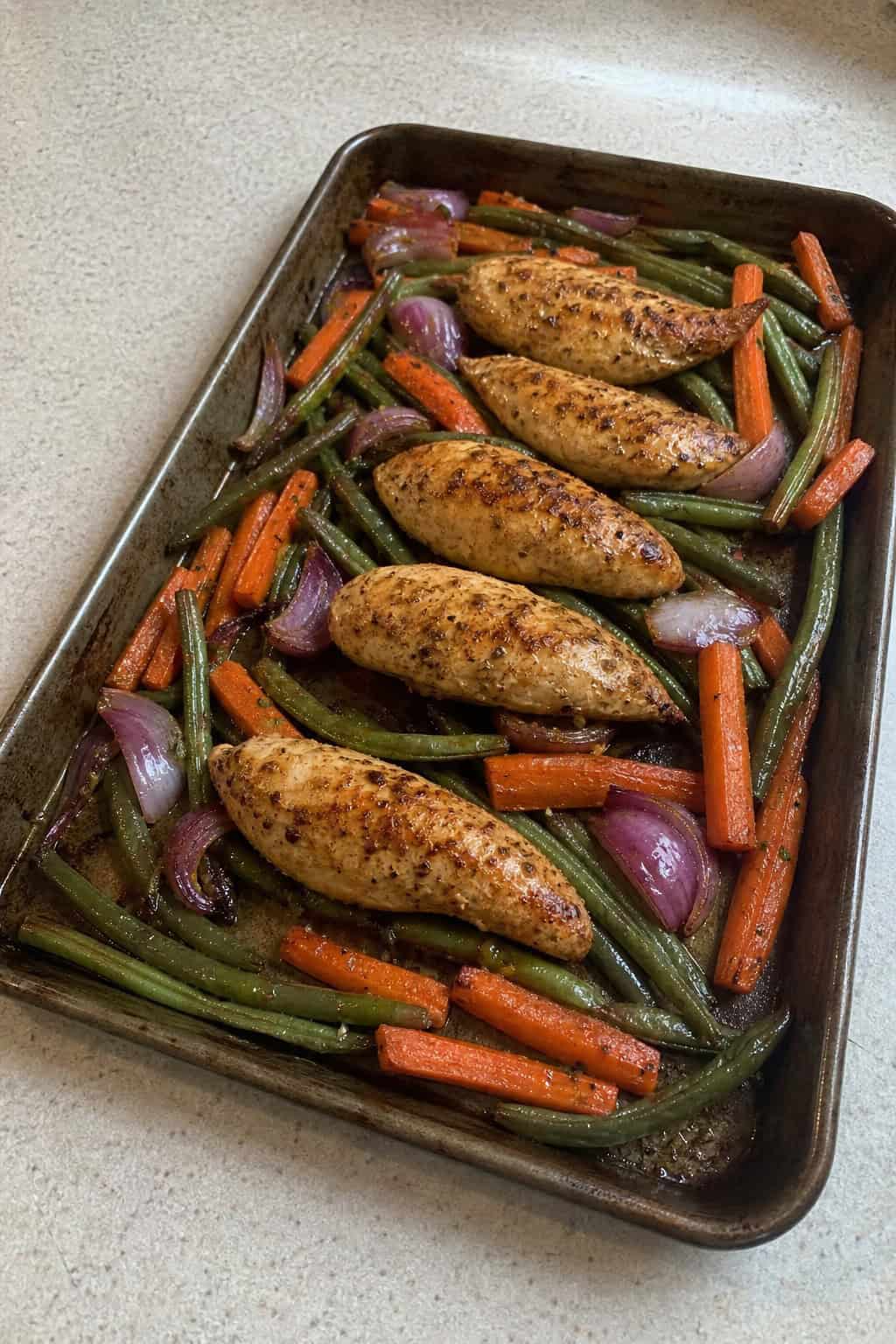 Sheet pan with golden chicken tenders and roasted vegetables on a kitchen countertop.