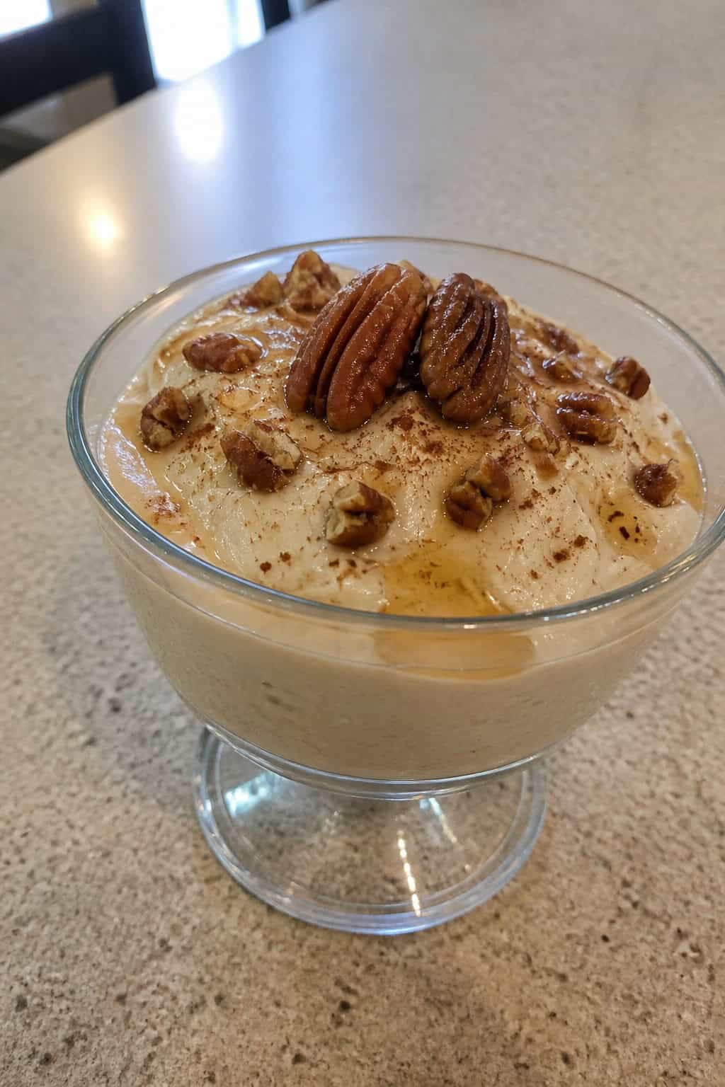 A glass cup filled with creamy maple pecan cheesecake pudding topped with pecans and syrup on a wooden table.
