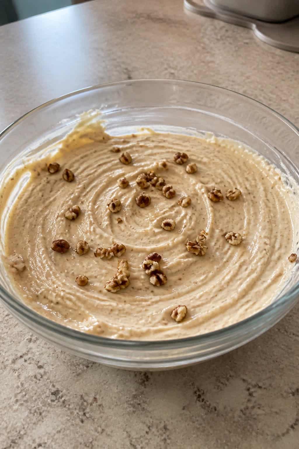 Close-up of creamy maple pecan cheesecake filling in a glass bowl with pecans and maple syrup on a kitchen countertop.
