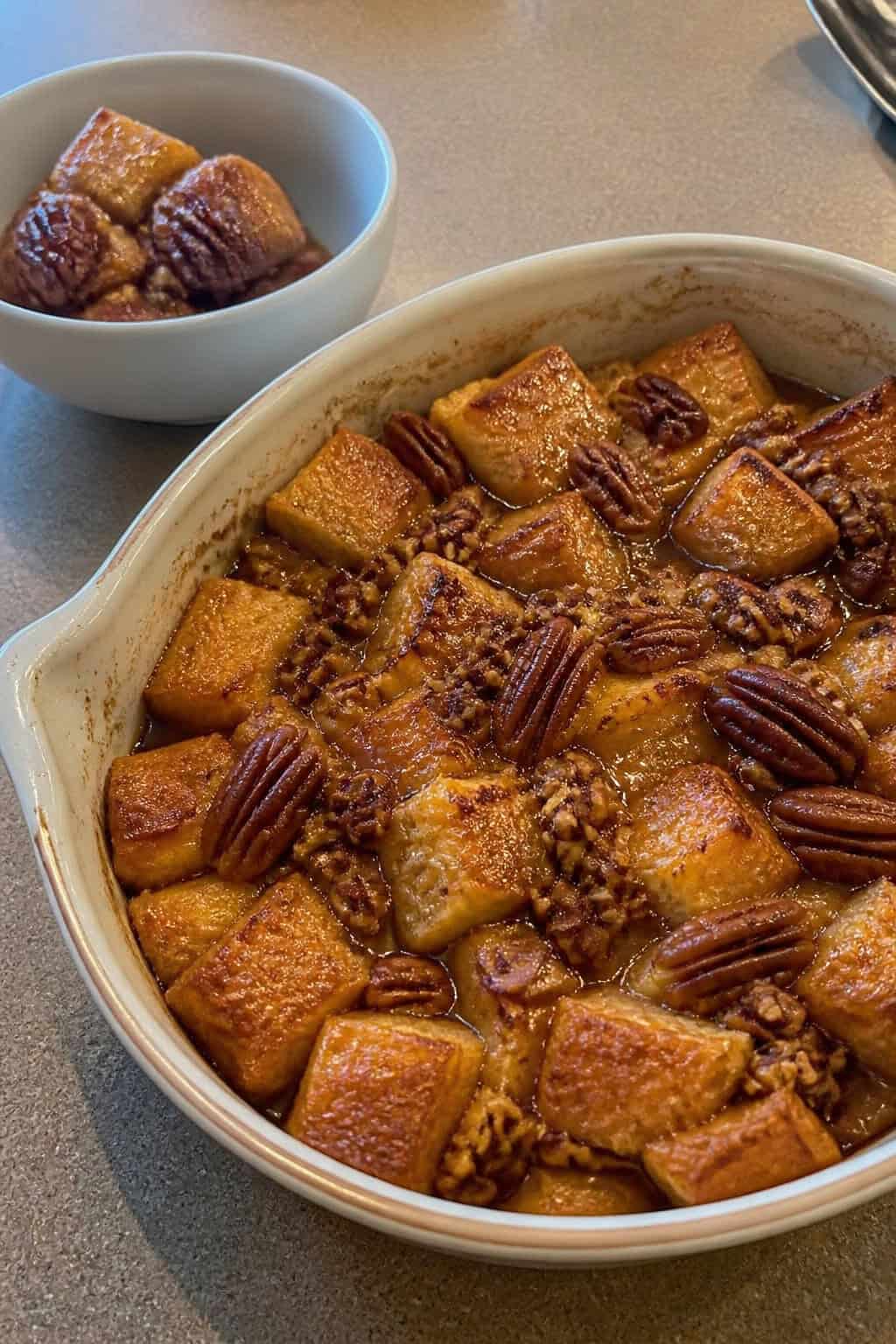 A golden-brown French toast casserole topped with whole and chopped pecans and a drizzle of maple syrup in a white ceramic dish on a wooden table, with a small jar of syrup and scattered pecans alongside.