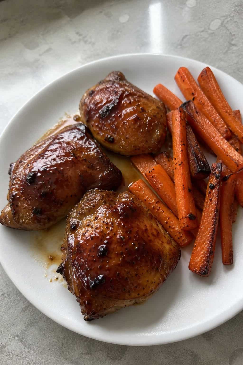 A plate of maple-soy glazed chicken thighs served with roasted carrots on a wooden table.