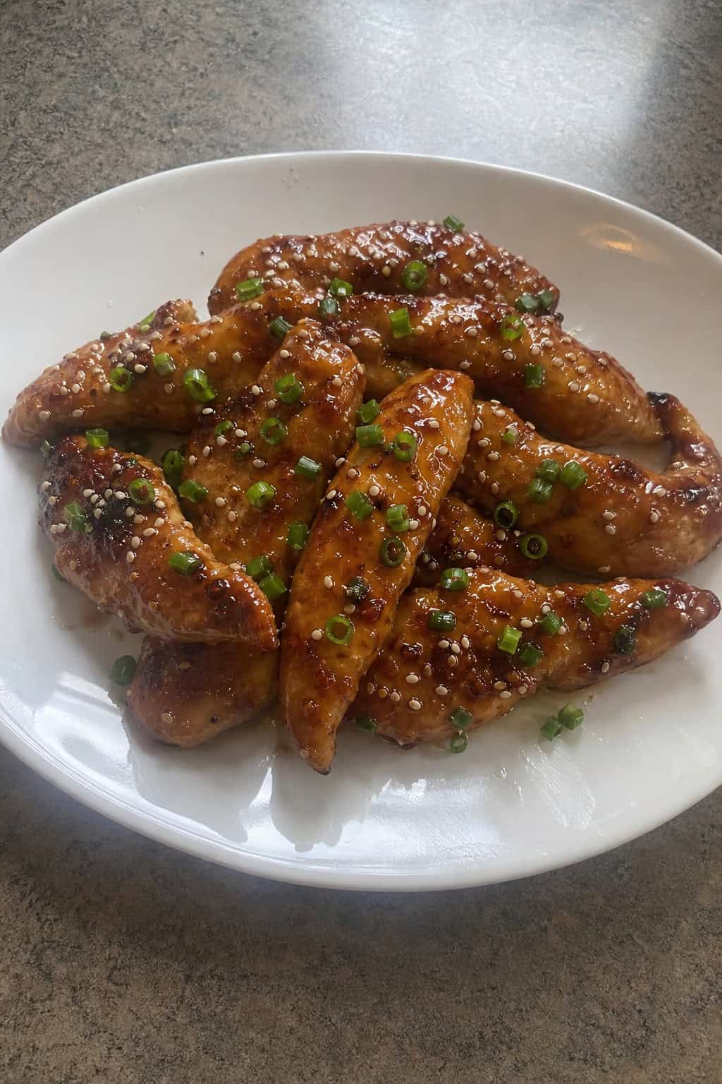 Glazed maple-soy chicken tenderloins topped with sesame seeds and sliced green onions on a wooden serving board.