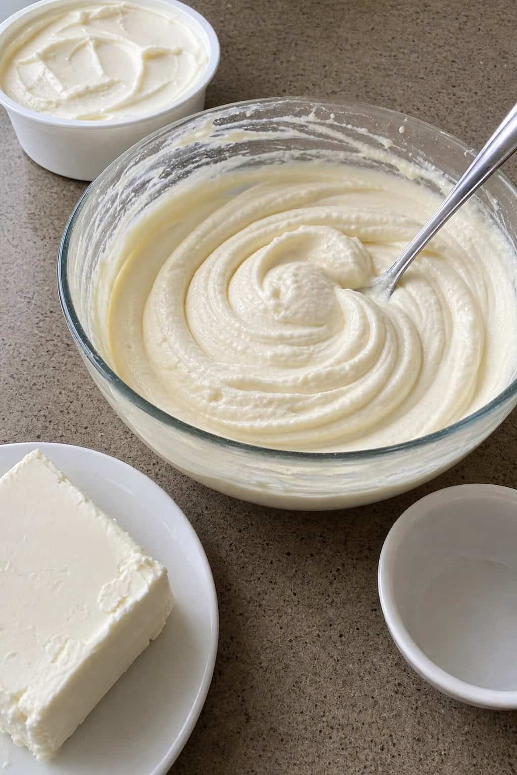 A glass bowl filled with smooth creamy cheesecake filling surrounded by cream cheese, mascarpone, vanilla bean, and sugar on a kitchen counter.