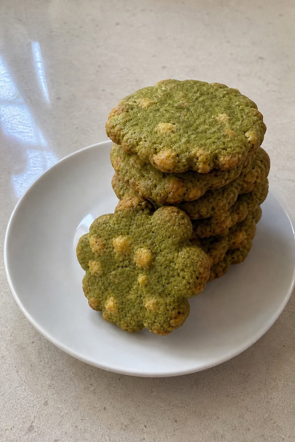 A plate of green matcha cheddar tea cookies on a wooden table with tea leaves, matcha powder, and cheddar cheese nearby.