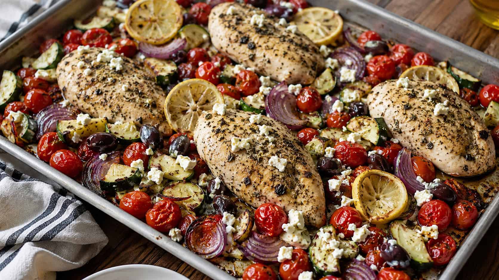 25 Heart-Healthy Chicken Recipes to Keep You Feeling Great! 28 a finished sheet pan Mediterranean chicken bake, roasted chicken breasts surrounded by burst cherry tomatoes, zucchini half-moons, red onion wedges, black olives, crumbled feta and lemon slices, warm natural light, casual home cooking