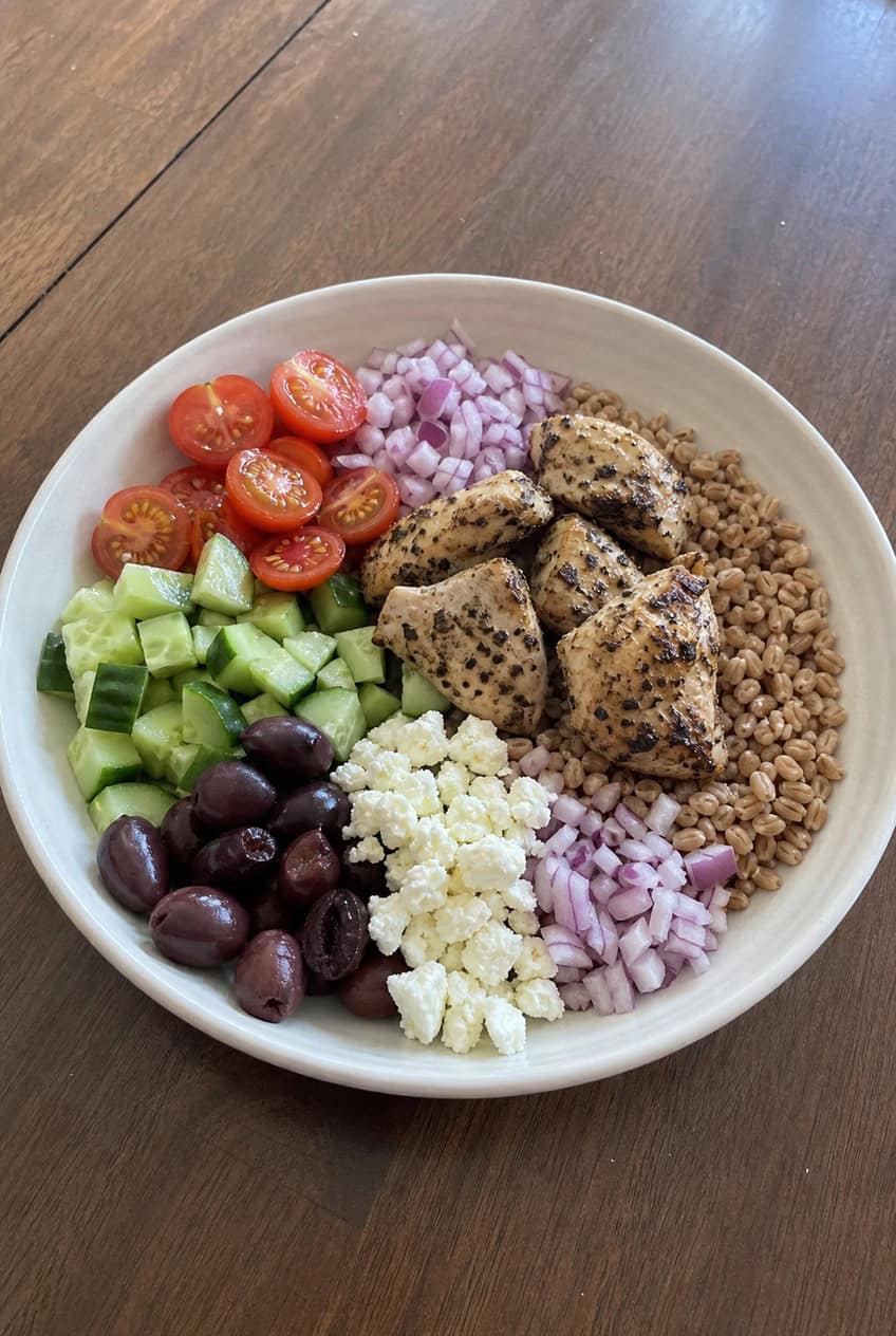 A wide grain bowl with a base of nutty farro topped with shredded rotisserie chicken, halved cherry tomatoes, diced cucumber, sliced kalamata olives, crumbled feta cheese, and thin-sliced red onion, drizzled with olive oil and lemon dressing, on a wooden table.