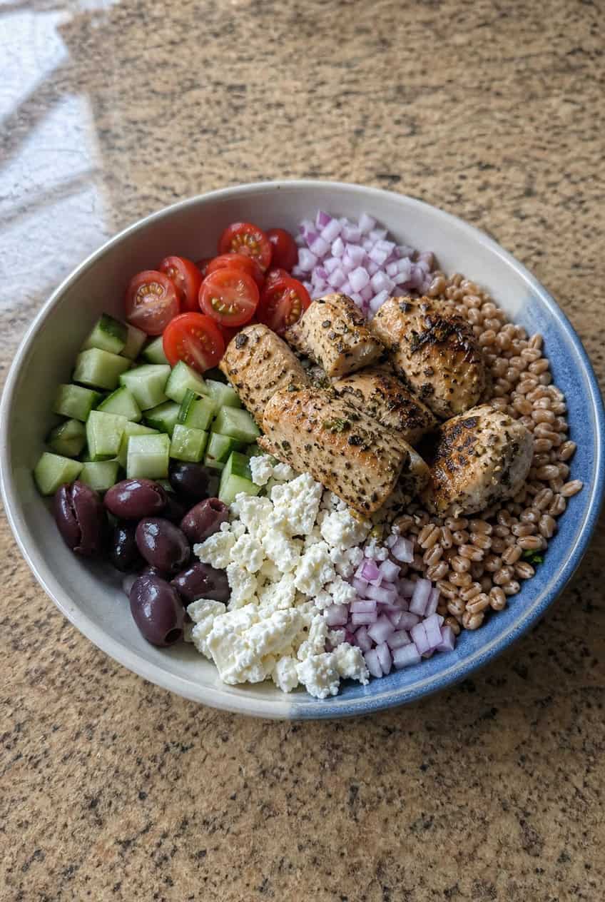 A wide grain bowl with a base of nutty farro topped with shredded rotisserie chicken, halved cherry tomatoes, diced cucumber, sliced kalamata olives, crumbled feta cheese, and thin-sliced red onion, drizzled with olive oil and lemon dressing, on a wooden table.