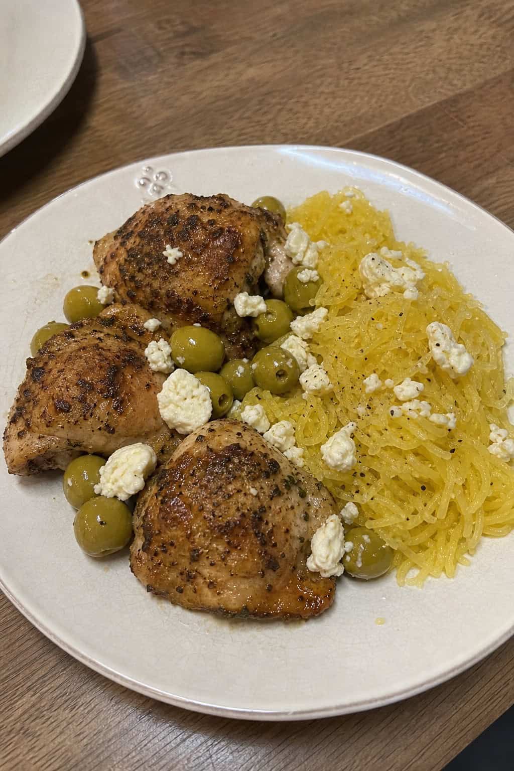 A plate of roasted chicken with olives, feta cheese, and spaghetti squash garnished with fresh herbs on a wooden table.