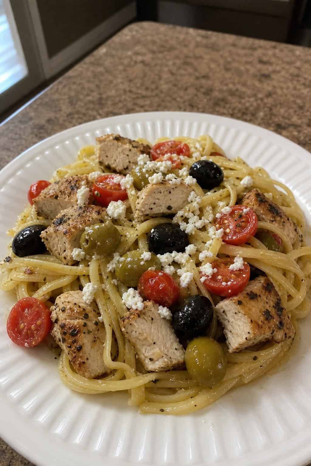 A bowl of Mediterranean chicken pasta with olives, feta cheese, cherry tomatoes, and fresh basil on a wooden table.