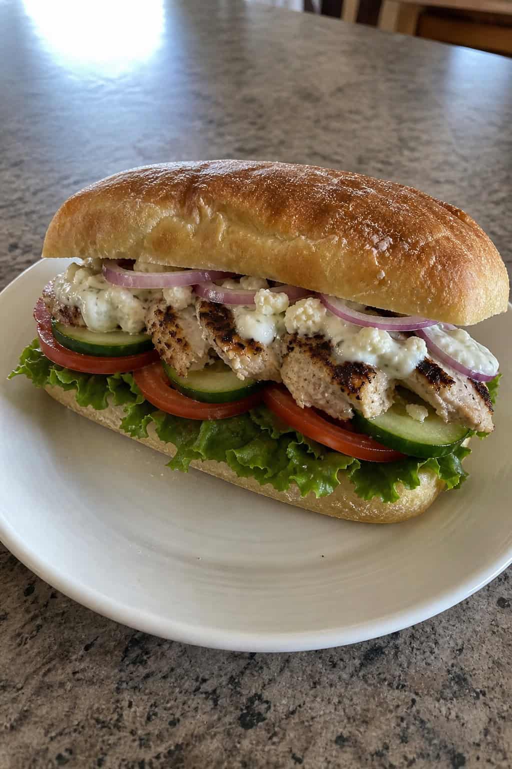 A grilled chicken sandwich with lettuce, tomato, red onion slices, tzatziki sauce, and crumbled feta on a toasted roll, surrounded by fresh ingredients on a wooden board
