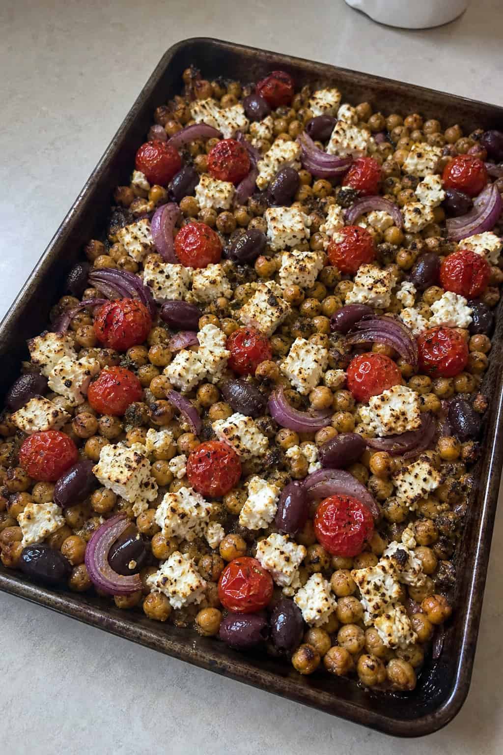 A foil-lined sheet pan filled with lightly crisped golden chickpeas, blistered cherry tomatoes, red onion wedges, sliced red bell pepper, and scattered black olives, topped with crumbled white feta and fresh herb leaves on a wooden table.