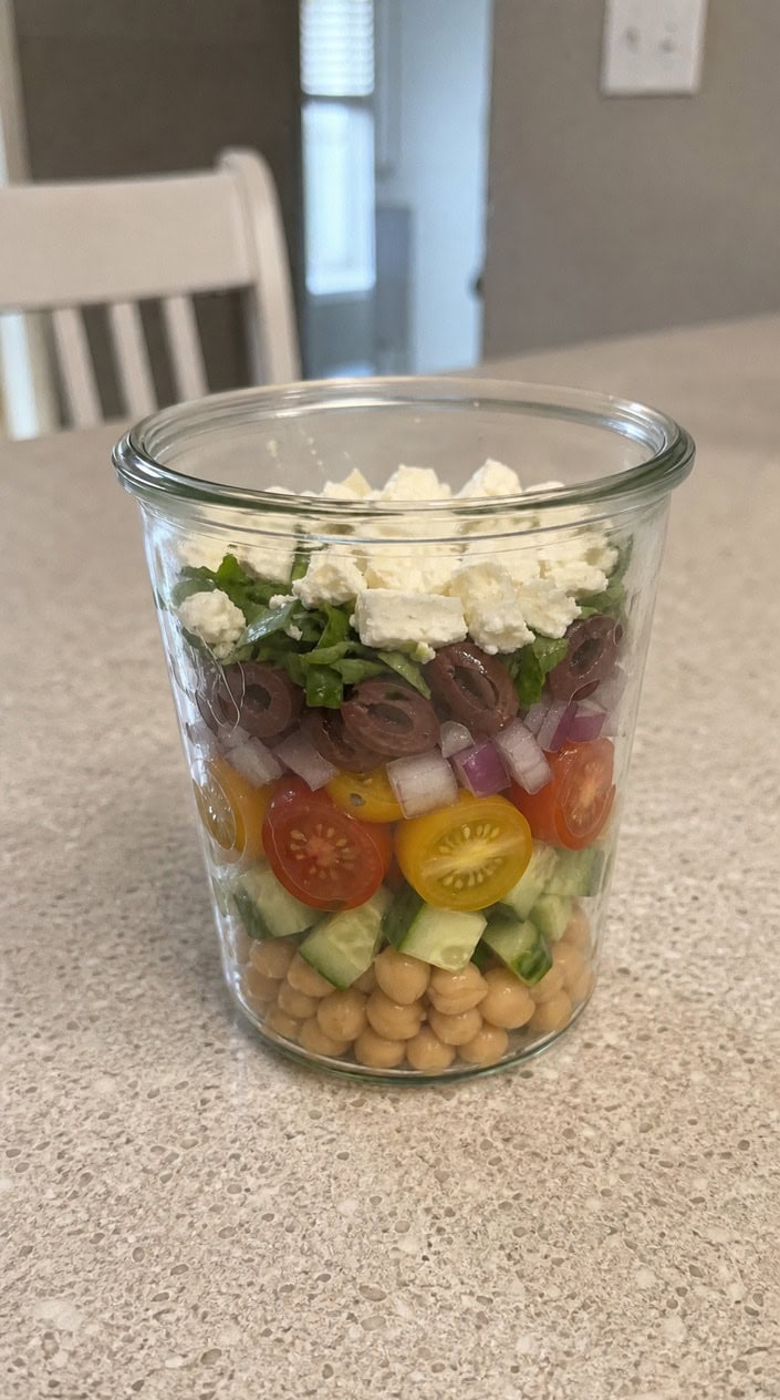 A wide-mouth glass jar layered with chickpeas, halved cherry tomatoes, diced cucumber, red onion, and crumbled feta, sitting on a warm-toned wooden surface with olive oil and a lemon half nearby.