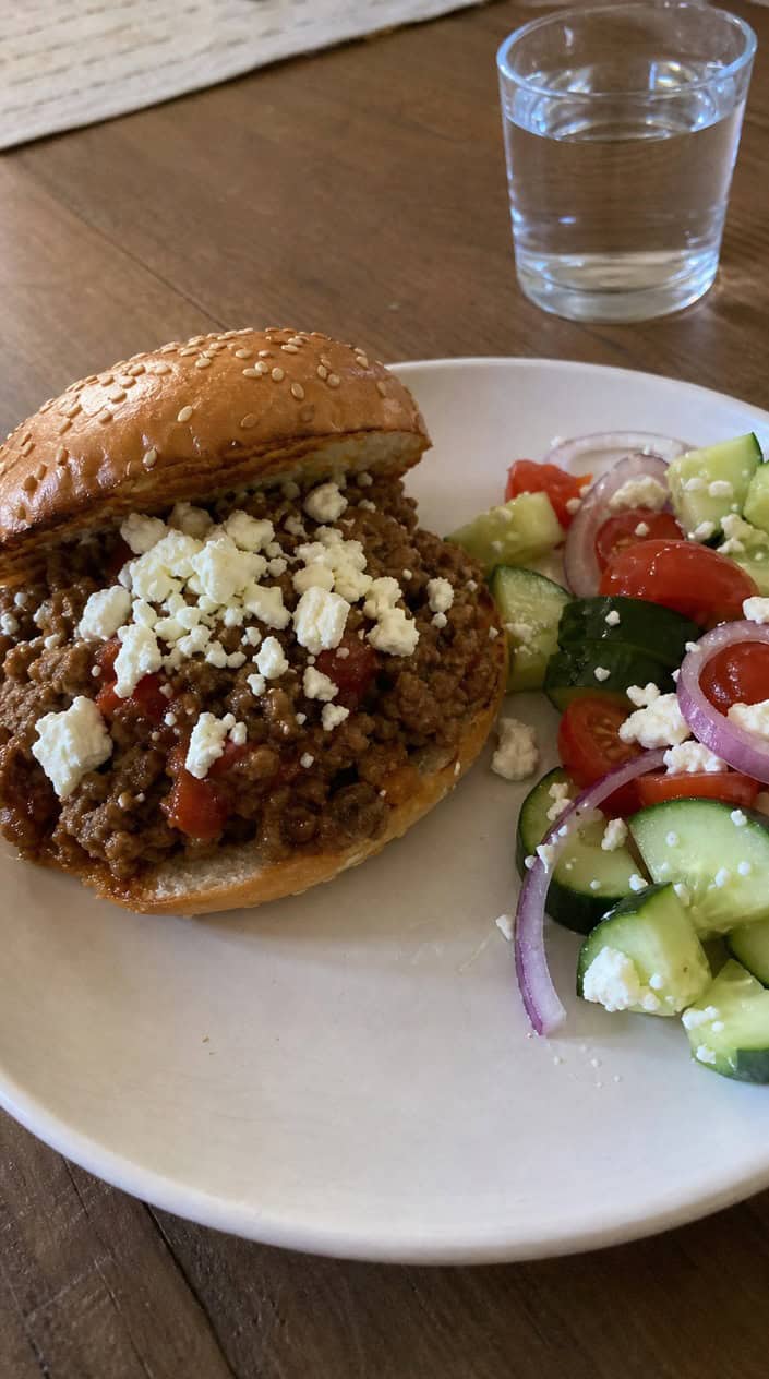 A lamb sloppy joe on a toasted roll, topped with crumbled white feta and bright green parsley leaves, with a small Greek salad visible on the plate beside it.