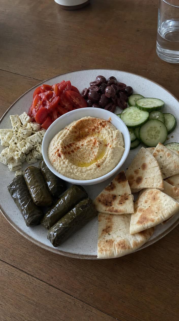 A Mediterranean mezze platter with hummus, dolmas, cucumbers, tomatoes, olives, and pita bread on a wooden table.