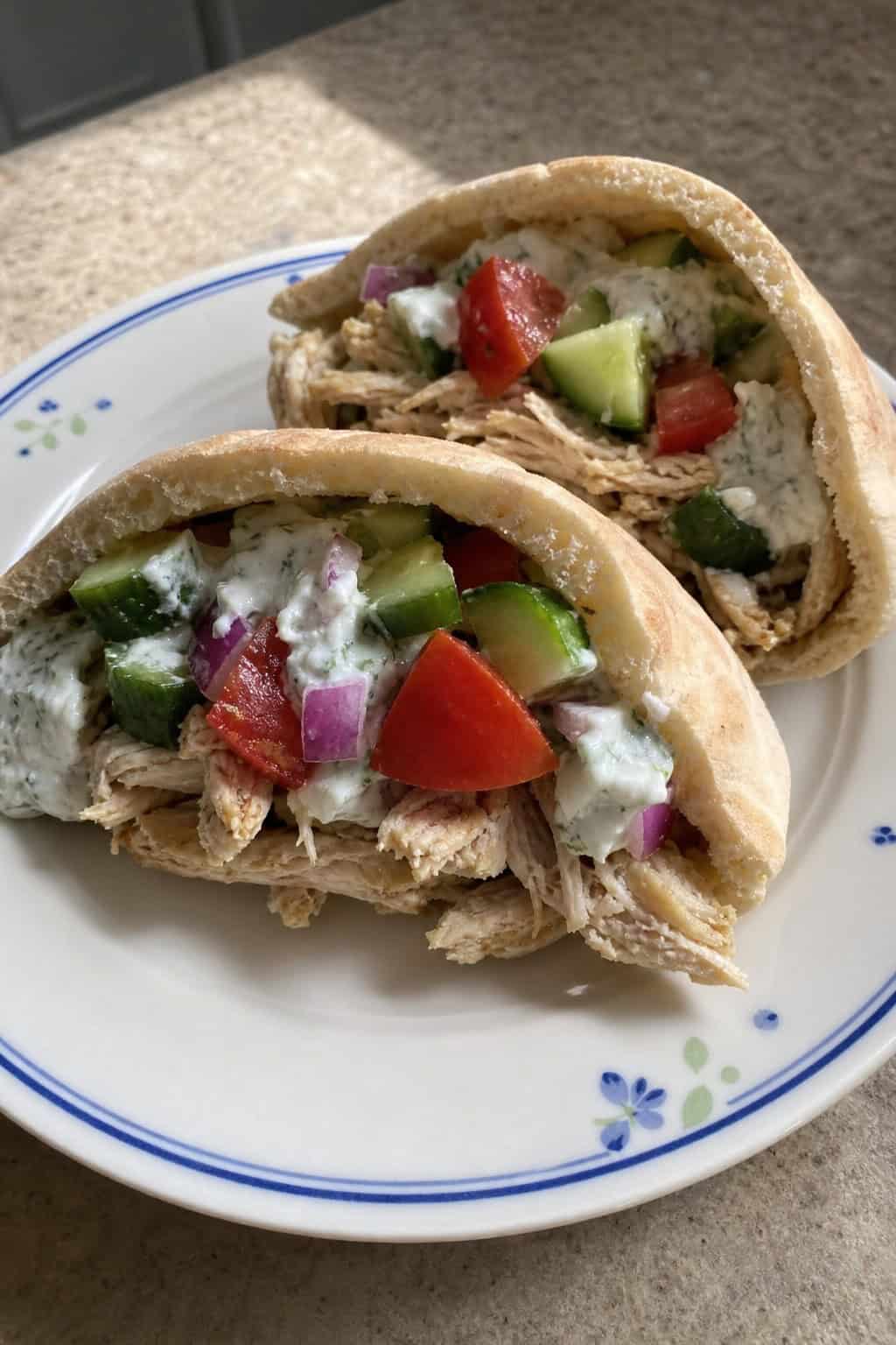 A wooden table with halved pita pockets filled with herbed shredded chicken, diced cucumber, halved cherry tomatoes, kalamata olives, and crumbled feta, with a small bowl of yogurt sauce alongside.