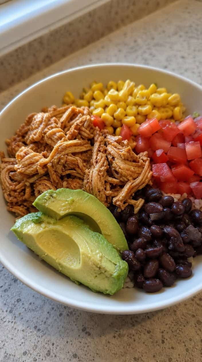 A bowl of Mexican chicken burrito bowl mix with chicken, beans, corn, tomatoes, avocado, and lime wedges on a wooden table.