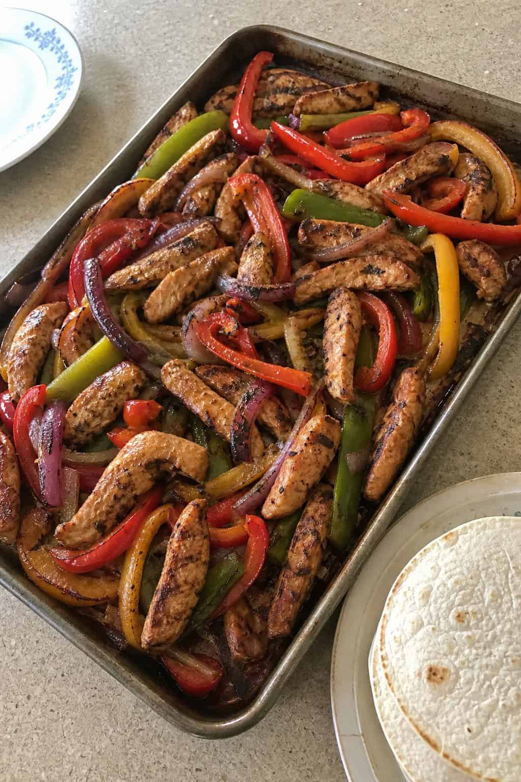 A foil-lined sheet pan with spice-coated sliced chicken thighs and charred bell pepper strips in red, yellow, and green, with crisp-edged tortilla wedges arranged along one side on a wooden surface.