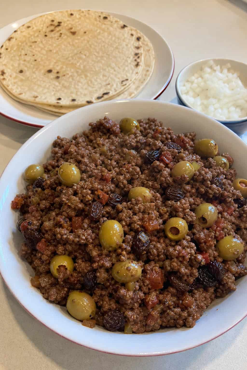 A bowl of Mexican picadillo with ground beef, raisins, and olives, served with corn tortillas and fresh garnishes on a wooden table.