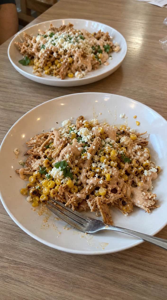 Generous bowls of seasoned shredded chicken over rice with bright yellow corn, fresh cilantro leaves, crumbled white cotija cheese, and lime wedges on a wooden table with fresh ingredients around the edges.