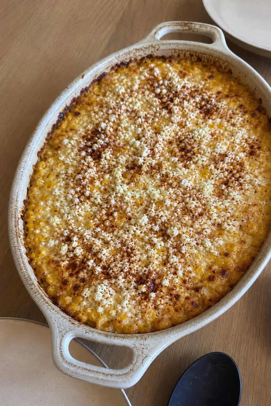 A golden baked corn casserole crumbled with white cotija cheese and scattered cilantro, surrounded by halved lime wedges and fresh jalapeños on a wooden table.