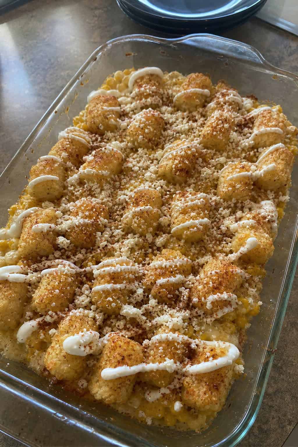 A baked Mexican street corn tater tot casserole in a ceramic dish with golden tots on top, garnished with bright green cilantro and lime wedges on the side, with small ingredient bowls arranged nearby on a wooden table.