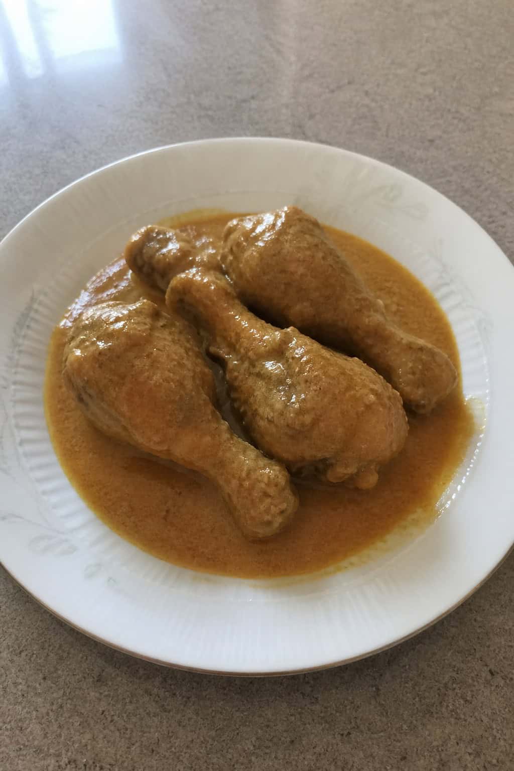 Lightly bronzed chicken drumsticks in a pale golden coconut curry sauce in a white baking dish, topped with fresh cilantro leaves, on a wooden surface.