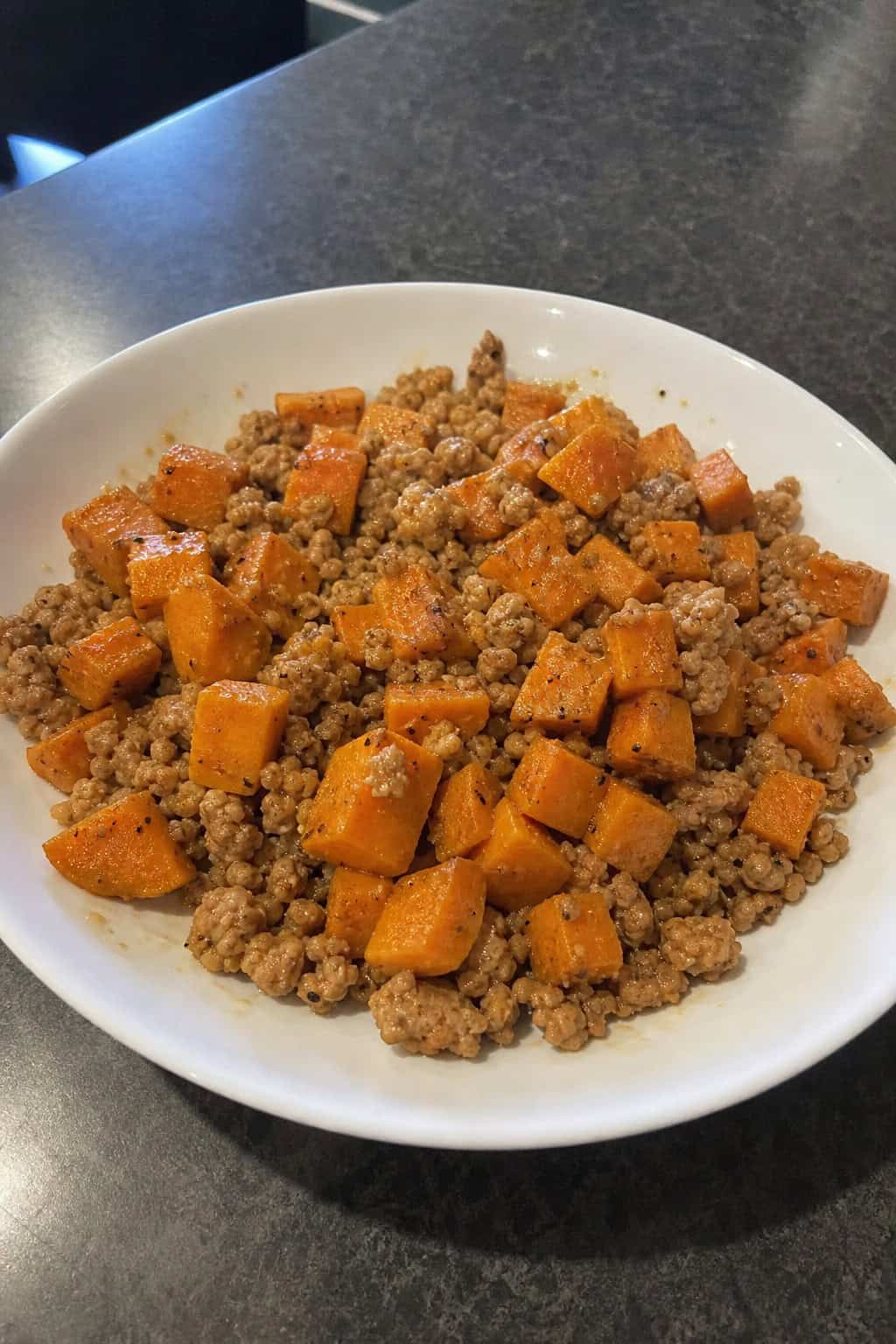 A bowl of minced chicken and sweet potato hash garnished with parsley on a wooden table with fresh ingredients around it.
