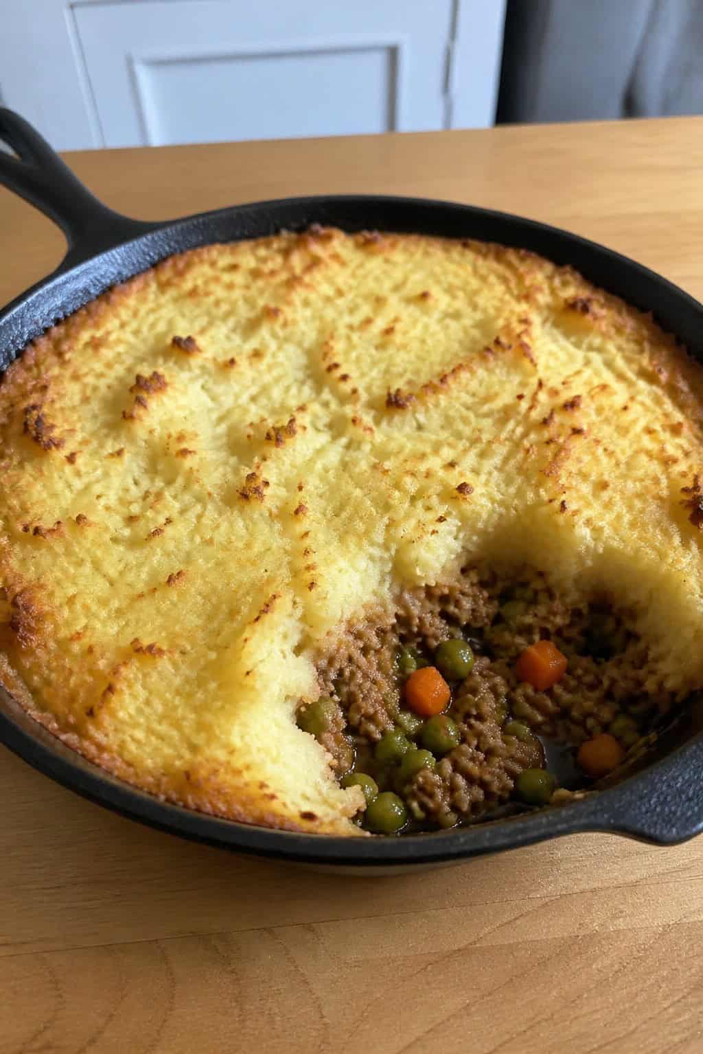 A skillet filled with minced chicken shepherd’s pie topped with golden mashed potatoes on a wooden table with fresh vegetables around it.