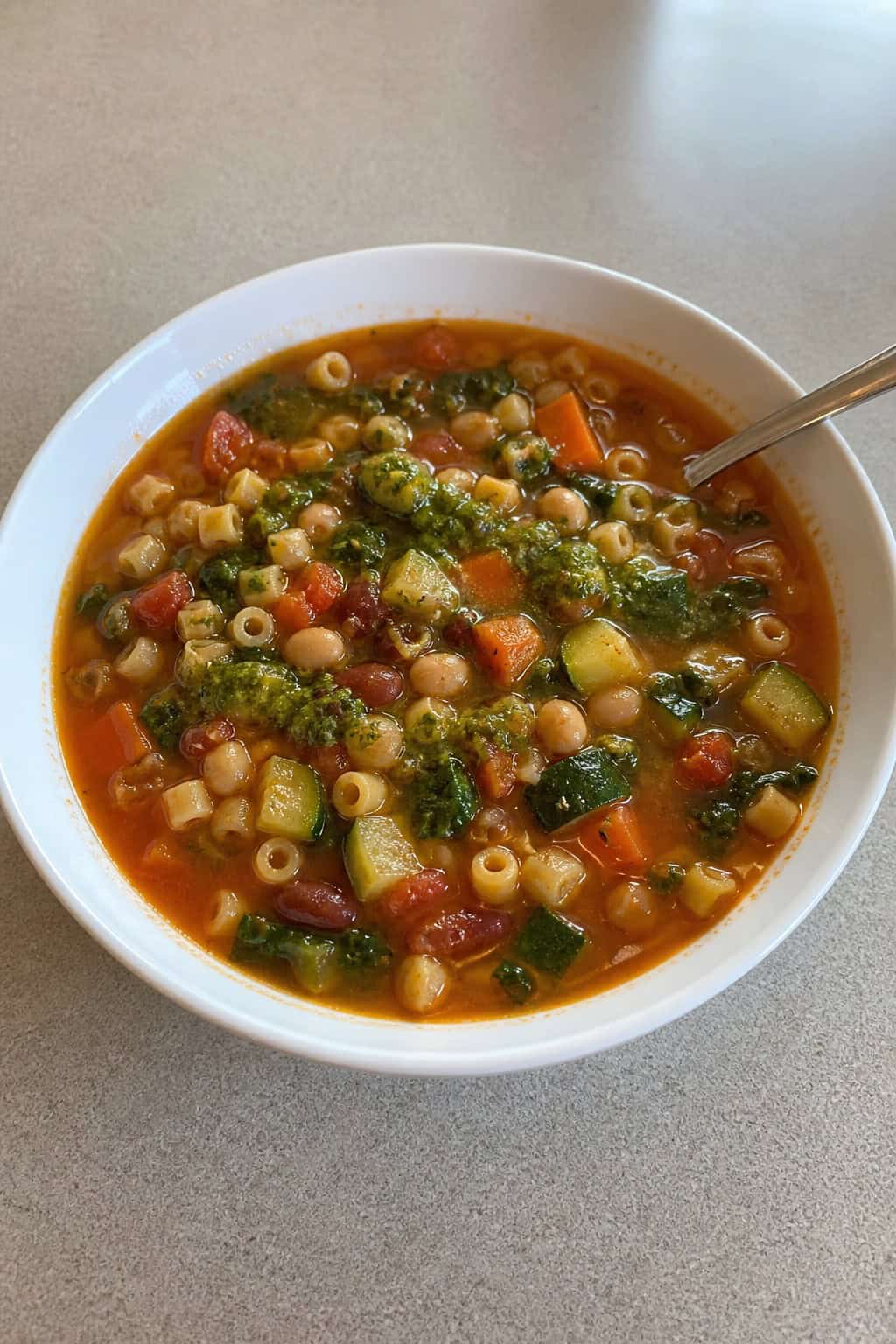 A bowl of minestrone soup with zucchini, pasta, and pesto on a wooden table with fresh ingredients around it.
