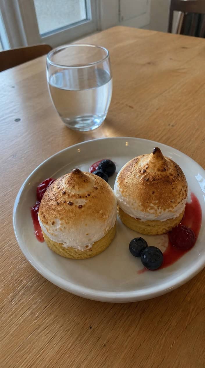 Two small Baked Alaska desserts plated on a white oval dish, each with a toasted golden meringue dome over a slice of pound cake, with two silver forks resting beside them on a softly lit table.