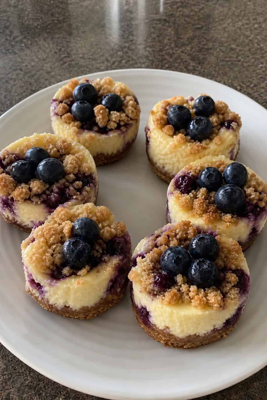 Several mini blueberry streusel cheesecakes on a wooden board with fresh blueberries around them.